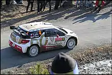 Vue plongée de la Citroën DS3 WRC de Sébastien Loeb, livrée blanche, rouge et or, sur une route goudronnée et entourée de spectateurs.