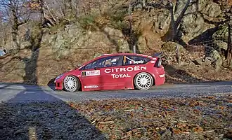 Photo de la Citroën C4 WRC de Sébastien Loeb, couleur rouge, vue de profil sur une route goudronnée avec un paysage montagneux.