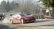 Citroën C4 WRC de Sébastien Loeb, livrée rouge, dans un virage sur une route goudronnée, foule de spectateurs en arrière-plan.