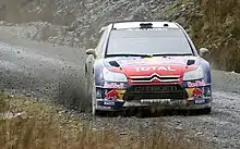 Citroën C4 WRC de Sébastien Loeb, livrée rouge et bleue, vue de face à pleine vitesse sur une piste boueuse.