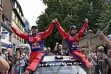Sébastien Loeb et son copilote Daniel Elena de face, assis sur le toit de leur Citroën C4 WRC, levant les bras au ciel et entourés de spectateurs.