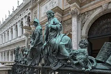 Image représentant la partie supérieure d'un portail surmonté de sculptures d'un chérubin au sol puis deux grands personnages, un deuxième chérubins au sol et à l'extrémité faisant face, un lion ailé tenant un livre ; en arrière-plan une grande façade de palais avec un grand portail en arc suivi d'arcades sur deux niveaux.