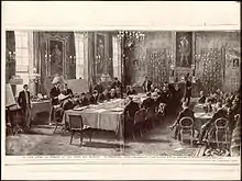photo noir et blanc d'une réunion d'hommes en costumes assis autour d'une longue table.