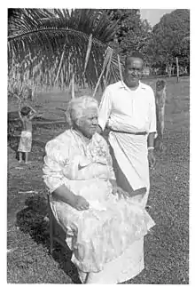Photo en noir et blanc de polynésiens habillés en blanc, une femme âgée assise et à ses côtés un homme debout