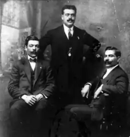 Trois hommes en costume sombre, portant moustache en guidon, cheveux gominés ; celui de gauche porte un nœud papillon, celui du milieu a une cravate et une montre à gousset, celui de droite porte une cravate.