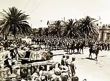 Des tankistes devant leur char et une foule regardent passer des cavaliers dans une rue bordée de palmiers.