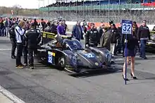 Photographie d'une voiture de sport-prototype, vue de trois-quarts, de couleur noire et or, sur la piste, entourée de personnes.