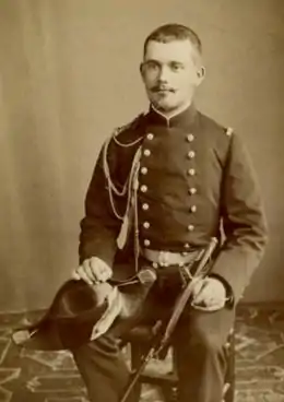 Photographie en noir et blanc d'un homme assis, portant une tenue de bordache, le poignard à la ceinture et le bicorne à la main.