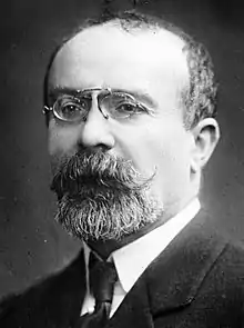Portrait en noir et blanc d'un homme en costume portant barbe et moustache et portant des lunettes rondes.