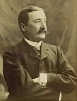 Photographie en noir et blanc d'un homme moustachu se tenant les bras croisés.