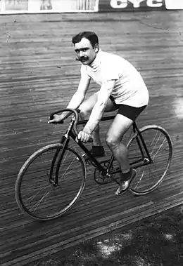 A black-and-white photograph of a man with three-colored sweater and shorts with a mustache sitting on a bicycle.