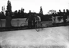 Photographie en noir et blanc montrant un cycliste en course, entrant sur la piste d'un vélodrome.
