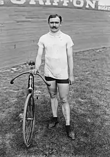 Photographie en noir et blanc d'un homme se tenant debout à côté de son vélo, au centre de la piste d'un vélodrome.