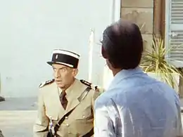 Entouré d'appareils de cinéma, un acteur habillé en gendarme joue la comédie, en étant observé par un homme de dos.