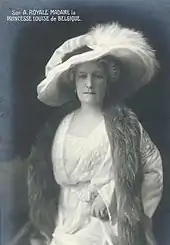 Photographie en noir et blanc d'une femme coiffée d'un grand chapeau à plume, elle porte une robe blanche et une fourrure autour de son cou.