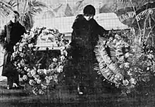 Louise Bryant, tenue sombre, le regard sur un cercueil à côté d'elle, couvert de fleurs