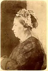 Photographie de Louise Milliet, de côté. Elle pose assise, habillée d'une robe, des fleurs dans les cheveux.