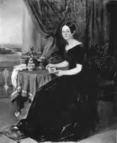 Portrait en pied de Louise Milliet mère, en robe, assise sur un fauteuil, les pieds posés sur un coussin à terre. Accoudée sur une table, elle tient un carnet entre ses mains. Plusieurs objets sont disposés sur la table.