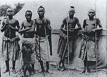 Photographie en noir et blanc présentant cinq Pygmées en costume traditionnel.