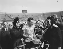 Photographie en noir et blanc d'un cycliste à l'arrivée d'une course, entouré par la foule et trois femmes en costume traditionnel breton.