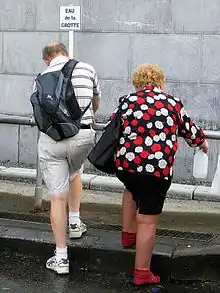 Photo en couleurs de deux adultes, remplissant chacun leur bouteille à un robinet, sous une pancarte marquée "eau de la grotte".