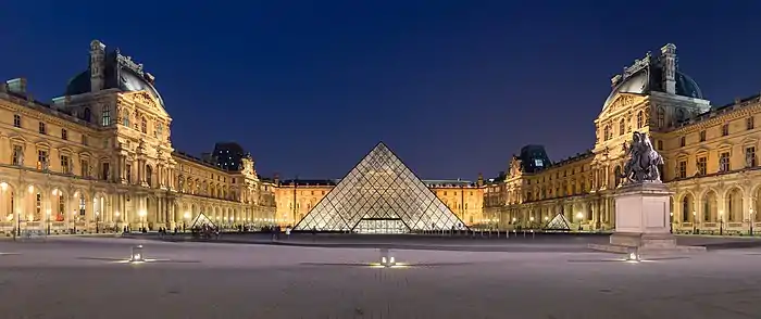 Une pyramide de verre se dresse la nuit au milieu d'une cour, tout autour des bâtiments plus anciens sont éclairés.