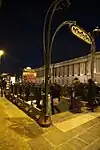 Photographie en couleurs la nuit d'une bouche de métro éclairée avec portique en fer forgé, longue façade illuminée sur la droite et ciel noir au-dessus