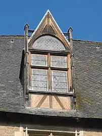 Photo couleur d'une fenêtre à meneau à encadrement de bois surmontée d'un fronton triangulaire percé d'un demi-cercle. Les ouvertures sont munies de vitraux à décor en losange.