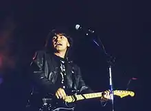 Photographie en couleur d'une personne sur scène, cheveux noirs, tenant une guitare et chantant, devant un haut micro sur pied