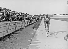 Photographie en noir et blanc d'un coureur cycliste roulant devant une foule de spectateurs groupés derrière une barrière en bois.
