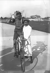 Photographie en noir et blanc d'un homme tenant le vélo d'un coureur cycliste à l'arrêt, qui tient un bouquet de fleurs d'une main et sur maintient sur l’épaule de l’homme de l’autre.