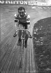 Photographie en noir et blanc d'un homme moustachu sur un vélo roulant sur un vélodrome.