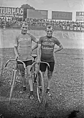 Photographie en noir et blanc de deux cyclistes debout, tenant leur vélo, dans un stade.