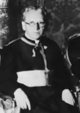 Photographie en noir et blanc du président du Zentrum, Ludwig Kaas, prise le 20 juillet 1933. L'image est floue et de très mauvaise qualité. Assis, Kaas est revêtu d'une soutane sur laquelle il porte une large ceinture de ton clair. De corpulence frêle, il a le visage mince, les cheveux gris et porte des lunettes rondes ; ses bras reposent sur les accoudoirs de son siège, les mains pendant vers le bas