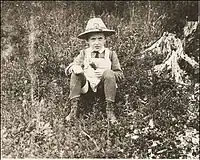 Photographie sépia d'un jeune enfant, assis de face dans les herbes hautes dans un champ fleuri.