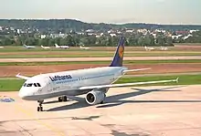 Photo granuleuse prise de trois quart face d'un avion de ligne aux couleurs blanches avec un logo jaune et bleu sur la queue, roulant sur le tarmac d'un aéroport.