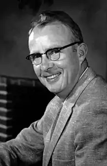 Photo en noir et blanc. Photo du torse d'un homme souriant qui porte un veston et des lunettes.
