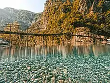 À travers une eau transparente, des cailloux bleus. Puis des reflets lumineux. Puis une passerelle fragile montée sur de hautes perches de bois ; puis une montagne arborée bouche la vue.