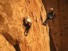 Une personne, accrochée, filmant une autre personne qui escalade la montagne.