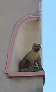 Statue, en hauteur, d'un chat assis dans une niche à l'angle d'un bâtiment. Vue selon son trois-quarts avant droit.