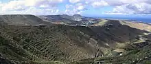 Paysage vallonné avec une végétation herbacée et arbustive et une ville dans la vallée. En arrière-plan, on distingue la mer.