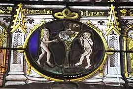 Médaillon de vitrail médiéval représentant la Tentation d'Adam et Eve, un homme et une femme sont nus autour d'un arbre portant des fruits et dans lequel il y a un serpent, la femme tient une pomme et l'homme a commencé à en manger une autre