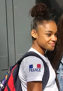 Portrait poitrine d'une jeune femme à la peau noire, les cheveux tirés et coiffés en chignon sur le dessus. Vue de profil, elle regarde vers la droite de la photographie et arbore un léger sourire. Elle est vêtue d'un t-shirt blanc dont la manche courte est ornée du drapeau français.