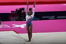 Une jeune femme en justaucorps blanc et bleu se tient debout sur un tapis, les jambes tendues et les bras levés le long des oreilles ; derrière elle, on aperçoit sur fond rose un logo blanc (celui de la compétition) ainsi qu'une caméra sur pied.