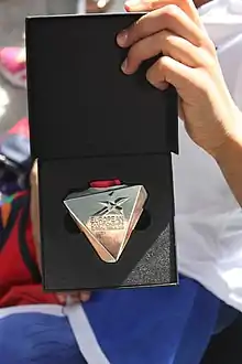 Une main tient ouvert un coffret noir dans lequel se trouve une médaille argentée de forme triangulaire, ornée d'un logo et du nom anglais de la compétition.