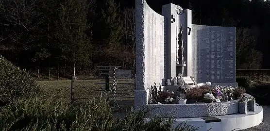 À droite, le monument aux morts en marbre ; derrière le monument, un grillage protège un tourniquet en métal.