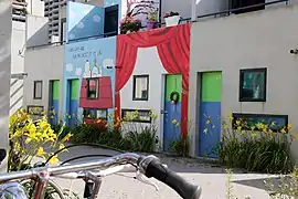 Quelques bungalows. Les portes sont peintes en bleu et vert et les murs sont décorés : un bungalow représente le personnage de bande dessinée Snoopy faisant la sieste sur sa niche, un autre représente des rideaux de théâtre. En avant-plan, le guidon d'un vélo. La rue est pavée et décorée de jonquilles de part et d'autre.