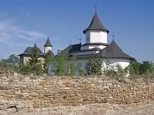 Mur en pierre derrière lequel se trouvent de la végétation et un bâtiment blanc au toit noir pointu.