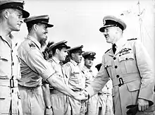 Deux hommes en uniformes militaires clairs avec des casquettes à visière se serrent la main devant une rangée d'hommes habillés de la même façon.