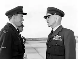Demi portrait informel de deux hommes portant des uniformes militaires sombres avec des casquettes à visière et qui tiennent une conversation à l'extérieur.